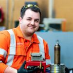 A young man in high vis overalls smiles at the camera as he works with engineering equipment.