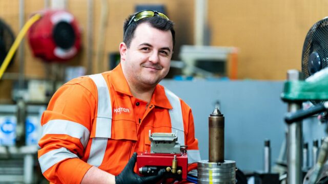 A young man in high vis overalls smiles at the camera as he works with engineering equipment.