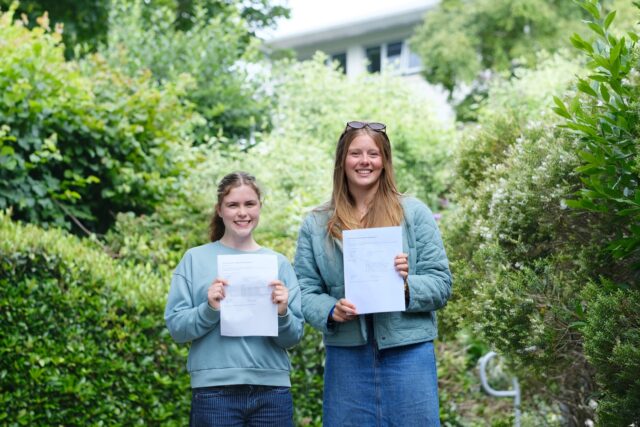 Bella and Lauren stand surrounded by greenery and hold their results in front of them.