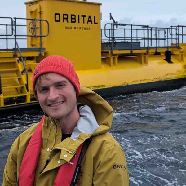 A man in a yellow waterproof coat with a lifejacket sits in front of large yellow machinery at sea. You can see the word 'Orbital, Marine Power' on his coat and on the machinery.