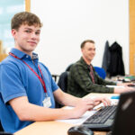An apprentice from the Eden Project sits at a computer with another apprentice in the background. He wears a blue shirt and smiles at the camera.