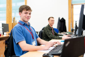 An apprentice from the Eden Project sits at a computer with another apprentice in the background. He wears a blue shirt and smiles at the camera.
