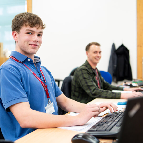 An apprentice from the Eden Project sits at a computer with another apprentice in the background. He wears a blue shirt and smiles at the camera.