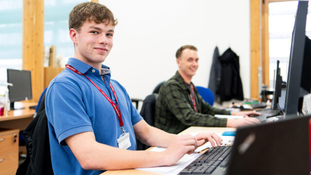 An apprentice from the Eden Project sits at a computer with another apprentice in the background. He wears a blue shirt and smiles at the camera.