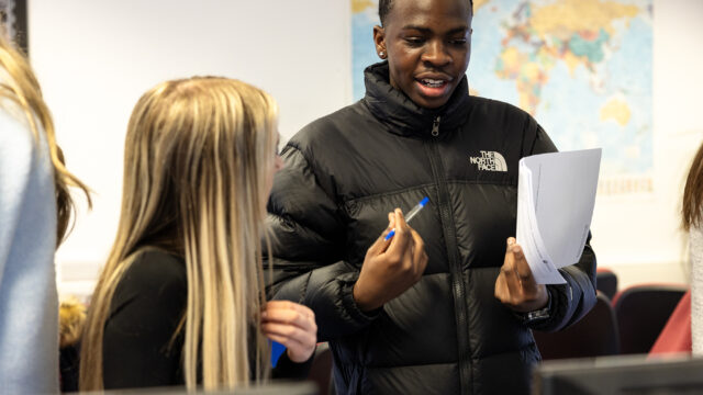 A student is talking to a member of staff. The student wears a dark coat and holds a pen in one hand and paper in the other.