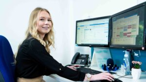 An apprentice sits at a computer with two screens open. There is a small plant pot on her desk and a phone. She wears a black top and a gold watch.