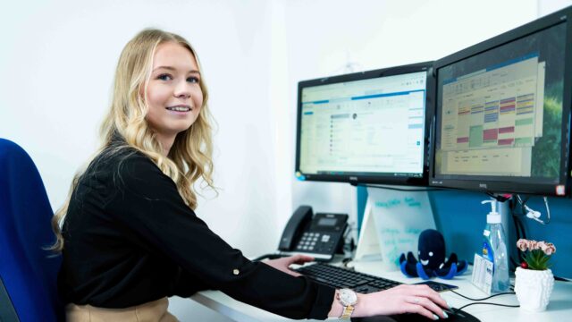 An apprentice sits at a computer with two screens open. There is a small plant pot on her desk and a phone. She wears a black top and a gold watch.