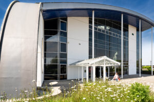 A picture of the outside of the Fal building at Truro College on a sunny day. There are grasses and daisies in the foreground.