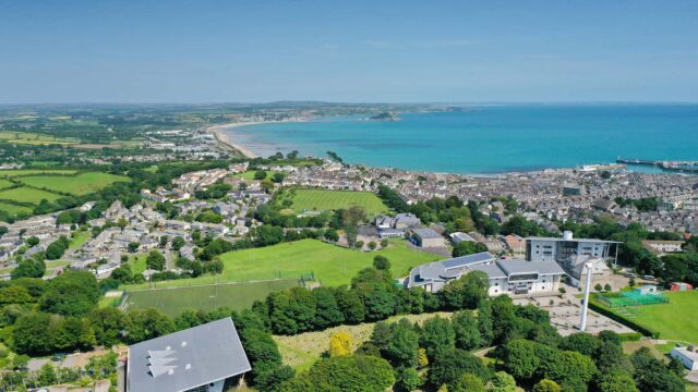 An aerial view over the Penwith campus out to sea and St Michaels Mount. There are lots of houses, fields and the sky is blue.