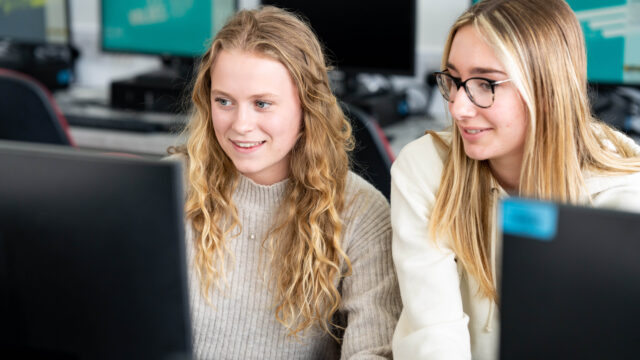 A photo of two girls looking at a computer screen together. They are both blonde and wearing cream jumpers and they are both smiling.