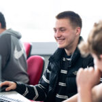 A boy sits at a computer smiling at the screen. He's wearing a stripey top and a body warmer.