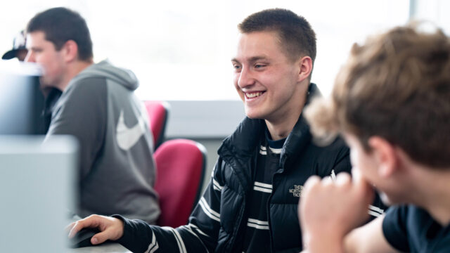 A boy sits at a computer smiling at the screen. He's wearing a stripey top and a body warmer.
