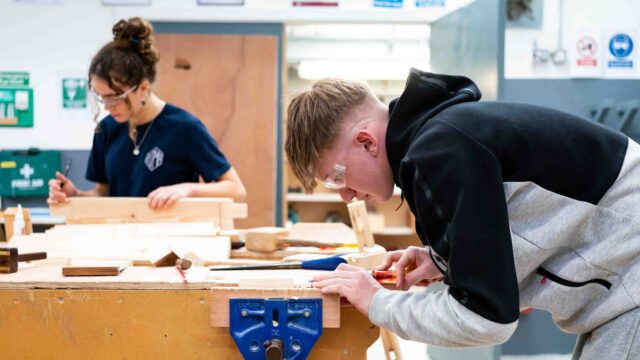 Two students work on wood in the construction area of the College.