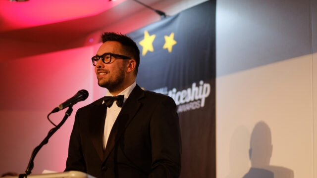 Neil Caddy stands at a lectern at the Cornwall Apprenticeship Awards. He is wearing a black suit and bow tie and has a banner up behind him.