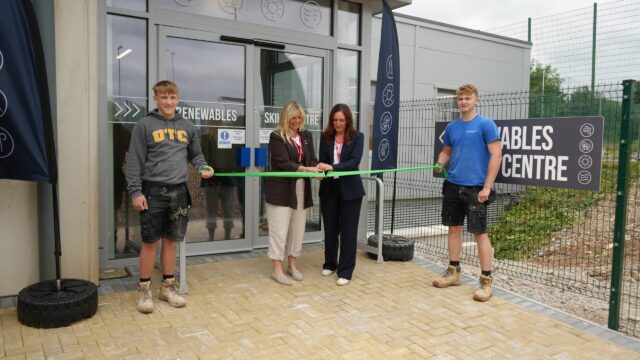 Two students hold out a green ribbon while two other people cut the ribbon with a pair of scissors.