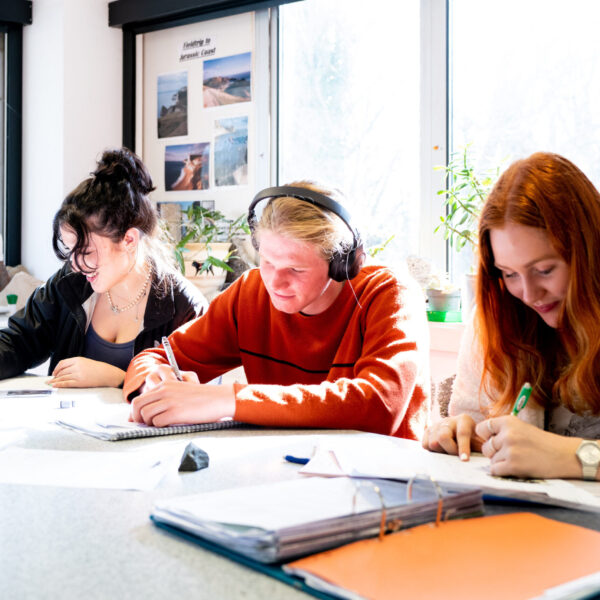 Three students sit at a table writing in notebooks.