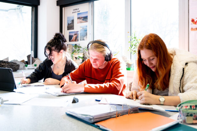 Three students sit at a table writing in notebooks.