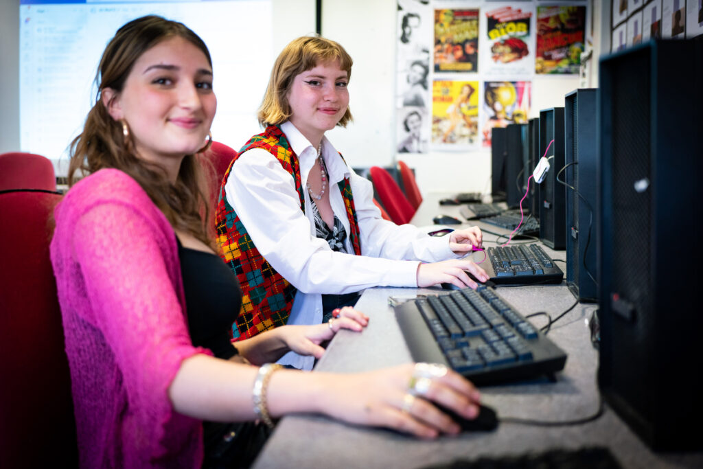 Two students sat at computers. There are media posters on the walls.