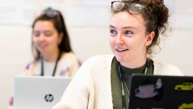 A close up of a student who has a laptop open in front of her. She wears a lanyard and has glasses on top of her head.