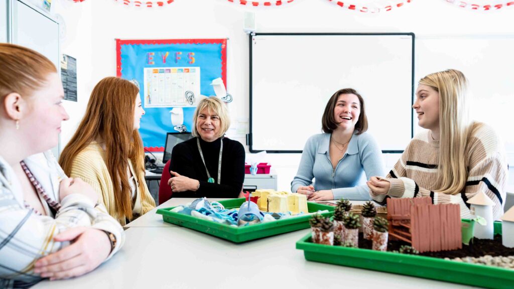 A group of students sit around a table and laugh with their tutor. There are craft items in front of them including pinecones and lolly-pop sticks.