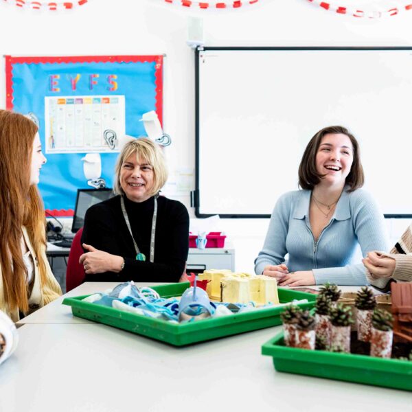 A group of students sit around a table and laugh with their tutor. There are craft items in front of them including pinecones and lolly-pop sticks.