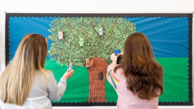 Two students pin child creations of birds and squirrels to a painting of a large tree.