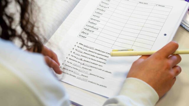 A close up image of a hand holding a pen and a workbook. The workbook has a table with spanish words, with gaps for the person to fill in.