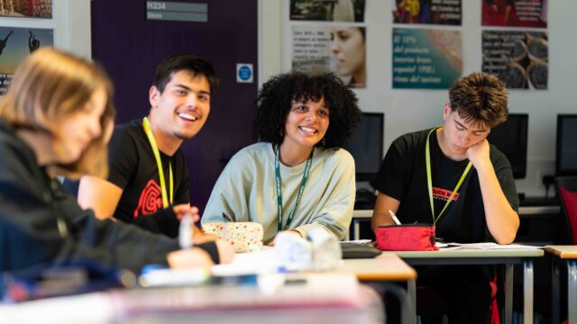 Four students sit at a table and look towards the front of the class. They are smiling and wearing lanyards.