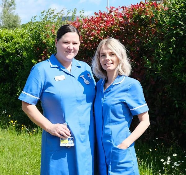 Two student nurses stand outside in the sun with their arms around each other.