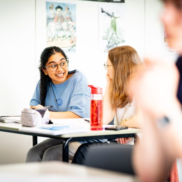 Two students sit at a classroom table chatting with water bottles and pencil cases on the table.