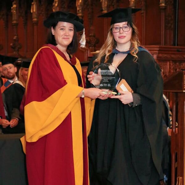 The Dean of the University Centre hands a trophy to student Scarlet. They are both in ceremonial gowns at the University Centre's graduation day.