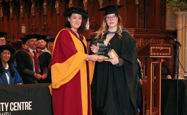 The Dean of the University Centre hands a trophy to student Scarlet. They are both in ceremonial gowns at the University Centre's graduation day.