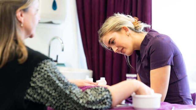 A hair and beauty student in a purple tunic is working on the nails of a client.