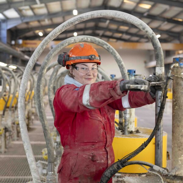 An apprentice in overalls holds a piece of equipment at arms length. She is wearing protective eyewear and a hard hat.