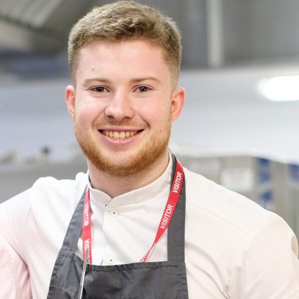 A headshot of Harrison. He is wearing a chef's apron and has a red visitor lanyard. He smiles at the camera.