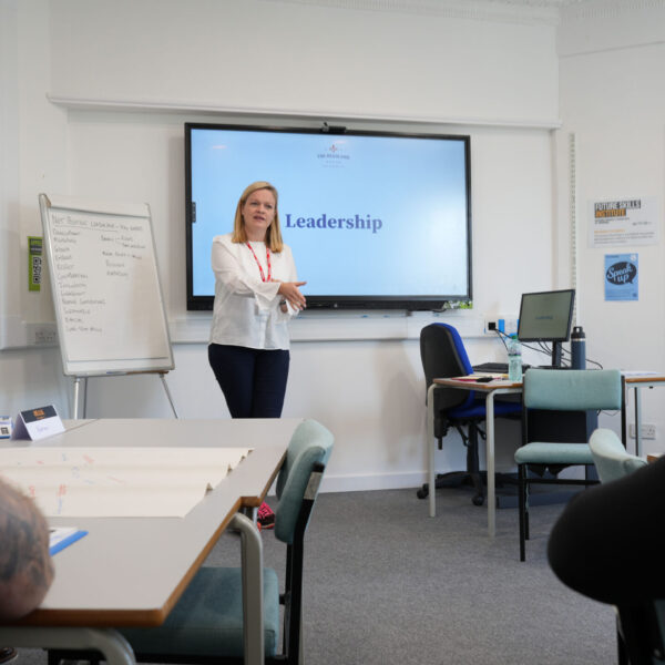 A tutor stands at the front of a classroom with the word 'leadership' on the board behind her.