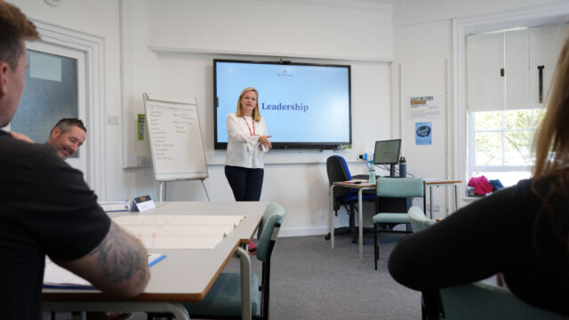 A tutor stands at the front of a classroom with the word 'leadership' on the board behind her.