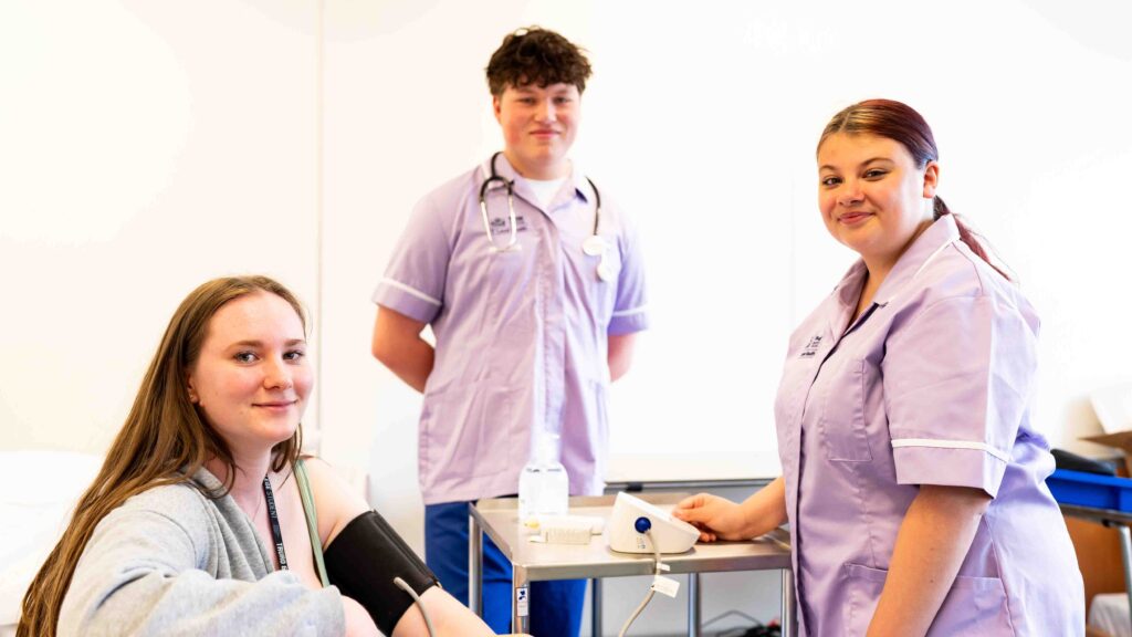 Three students look at the camera. Two are dressed in purple scrubs and the other has a blood pressure kit strapped to her arm.
