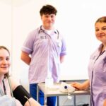 Three students look at the camera. Two are dressed in purple scrubs and the other has a blood pressure kit strapped to her arm.