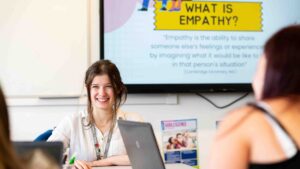 A tutor looks towards her students and smiles. She is sat at a table and the screen behind her says 'What is empathy?'