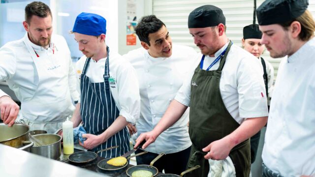Students work in a kitchen with chef Paul Ainsworth. They are using small frying pans on a large stove.