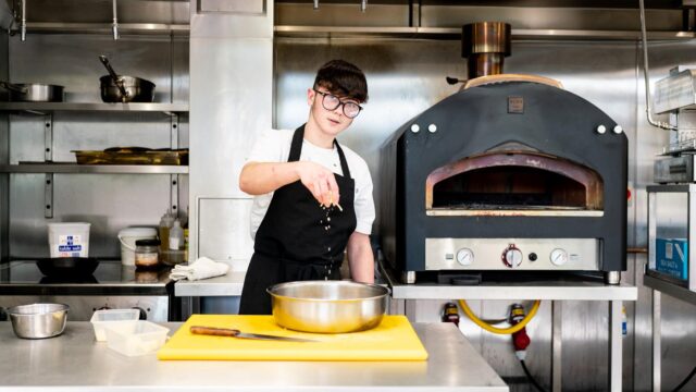A young boy dressed in an apron looks at the camera as he sprinkles cheese into a large stainless steel cooking bowl.