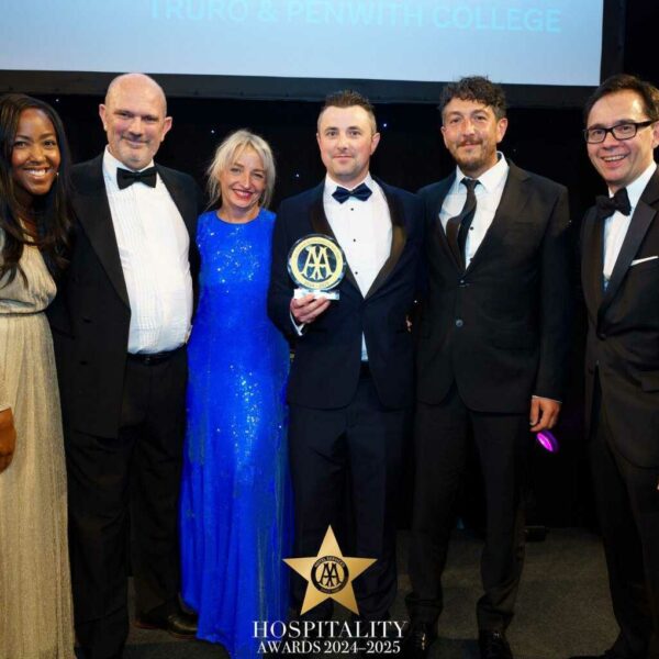 A row of 6 smartly dressed people have their arms around each other and the person in the middle holds up an award.