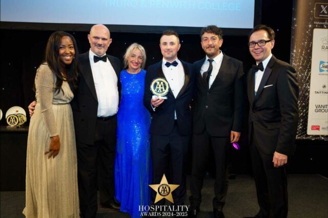 A row of 6 smartly dressed people have their arms around each other and the person in the middle holds up an award.