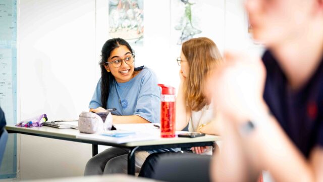 Two students sit at a classroom table and are chatting. They have pencil cases, notebooks and water bottles in front of them.