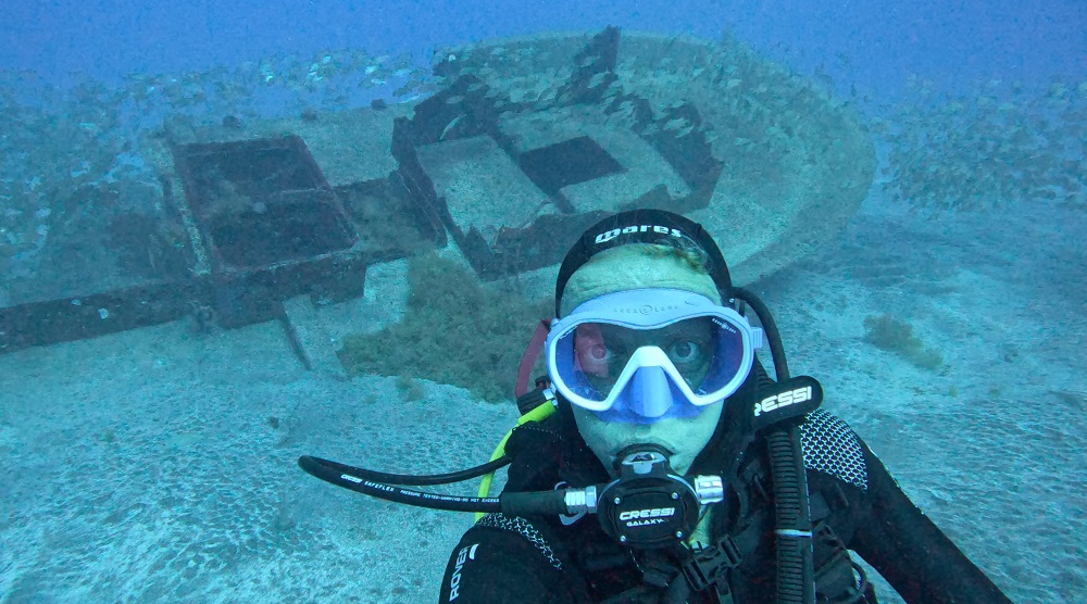 A selfie taken by James McBlane shows him underwater in full scuba diving gear with a shipwreck on the seabed behind him.