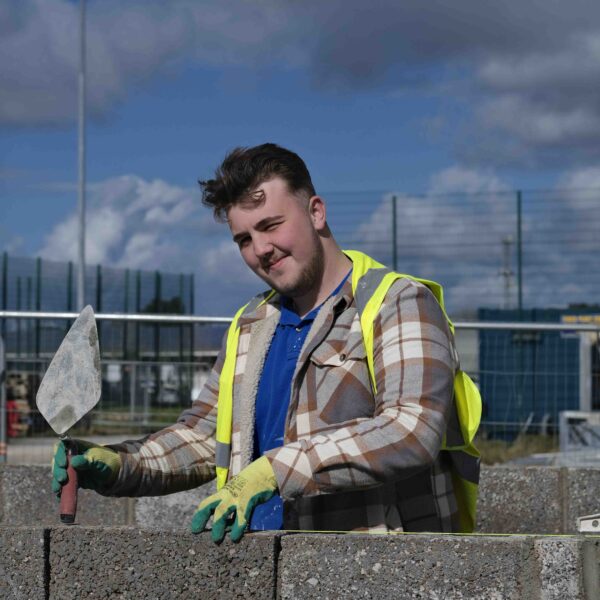 A student stands with a brick wall in front of them. They are holding a trowel and wearing a high vis jacket. It is a sunny day.