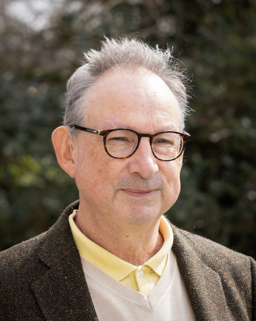 A portrait photo of Ken Curtis. He is wearing a yellow shirt, beige jumper and brown jacket.