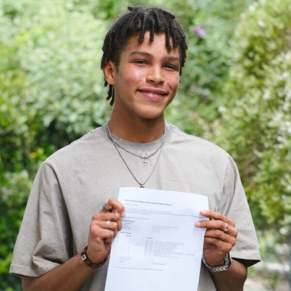Student, Louis, holds his results to the camera and smiles. He wears a grey shirt and there is greenery around him.