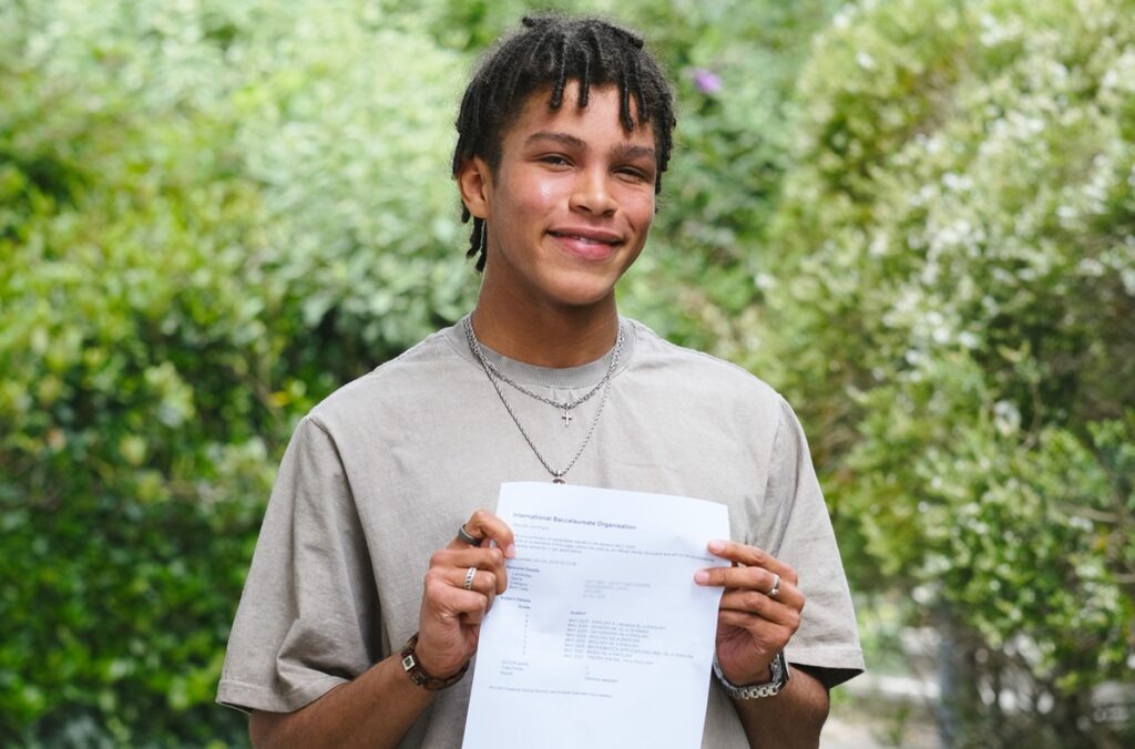 Student, Louis, holds his results to the camera and smiles. He wears a grey shirt and there is greenery around him.